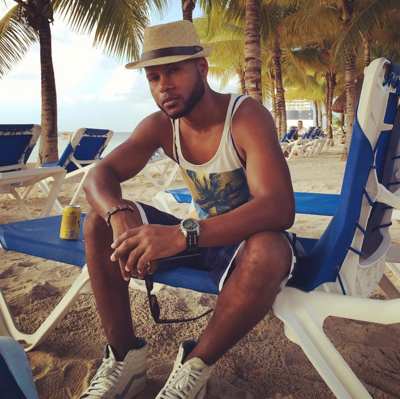 Anthony Alexander wearing a straw hat, tank top and shorts while seated beneath palm trees