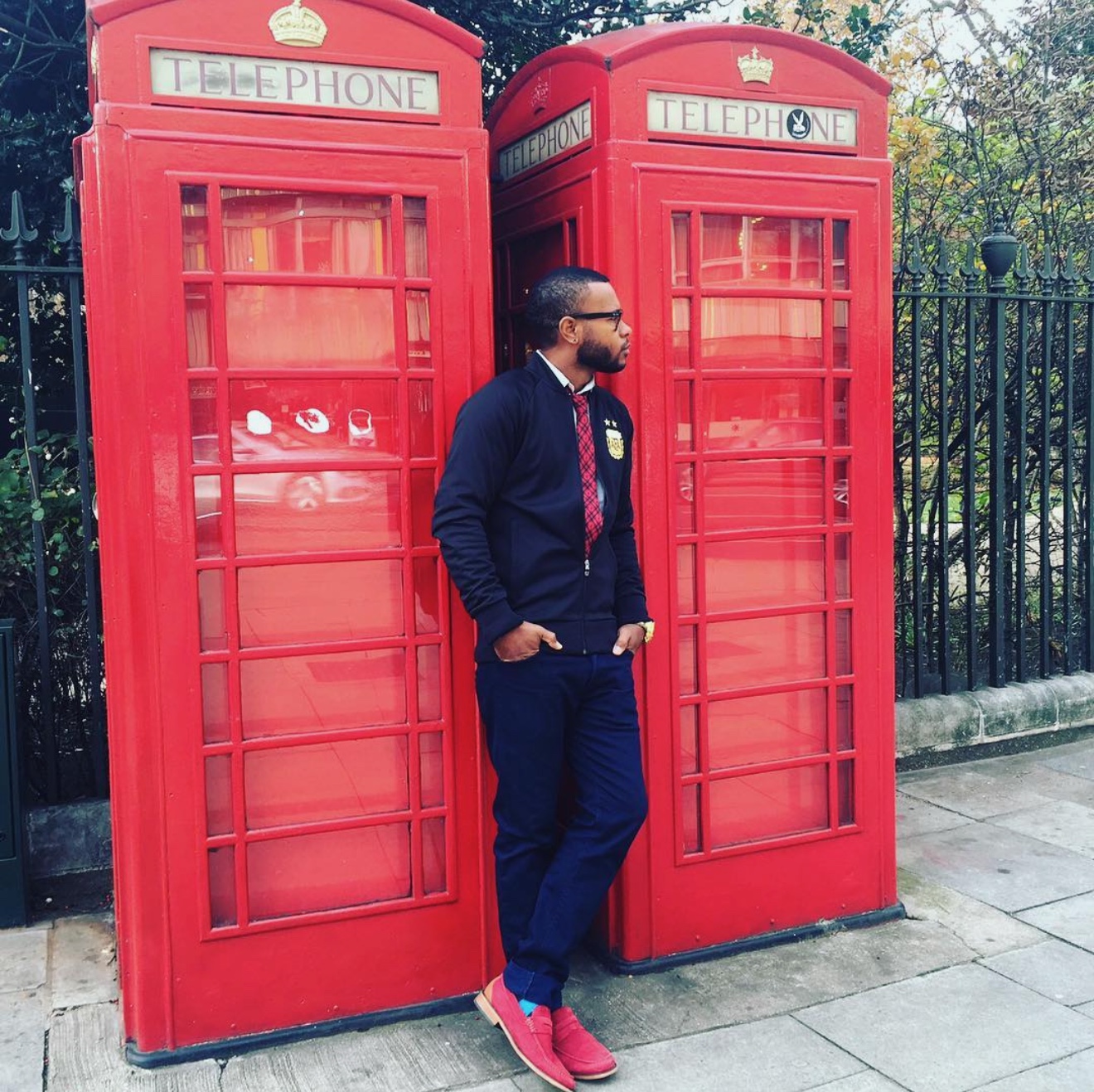 Anthony Alexander in a navy outfit and red loafers beside red telephone boxes