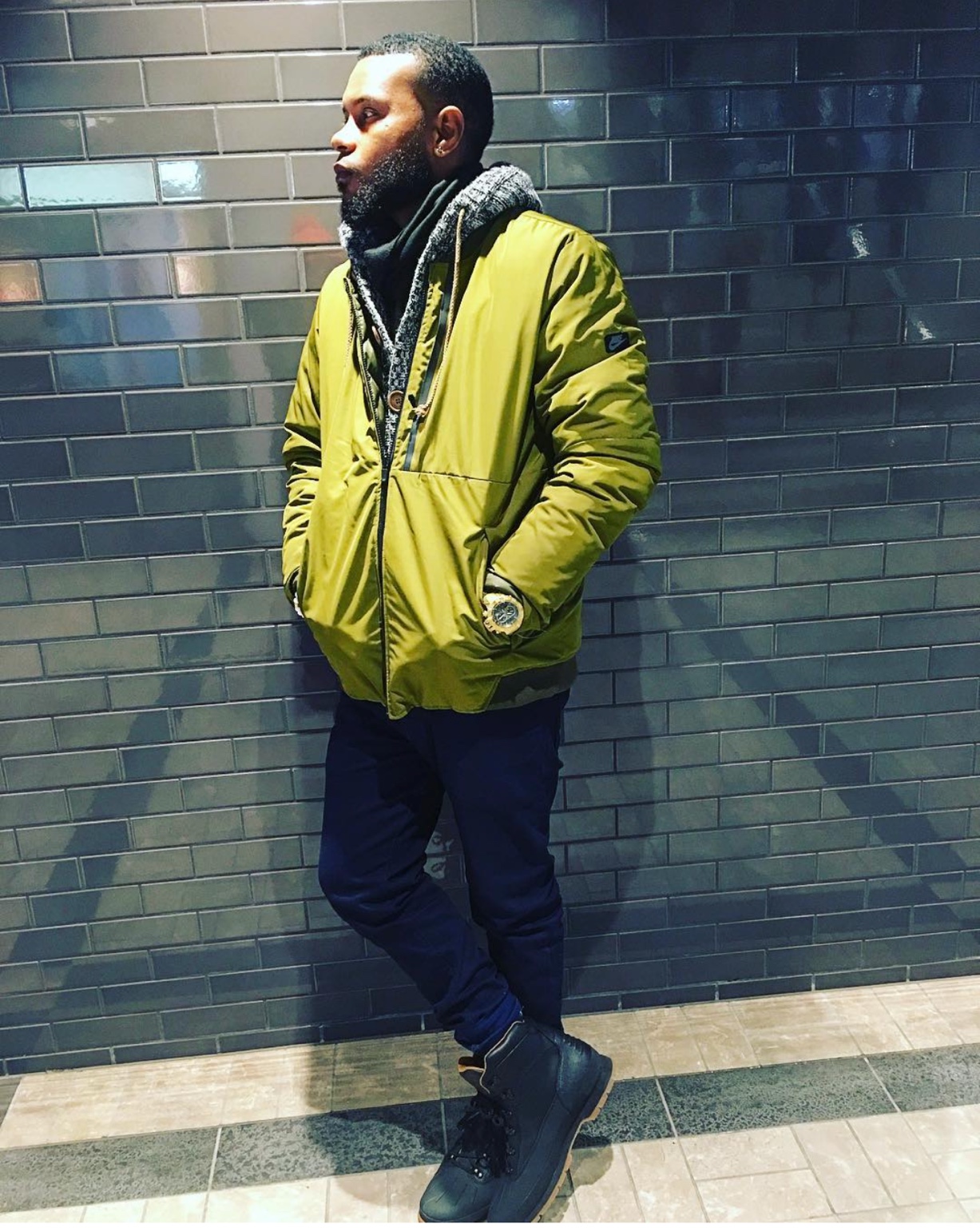 Anthony Alexander in an olive jacket, scarf, dark jeans and boots against a tiled wall
